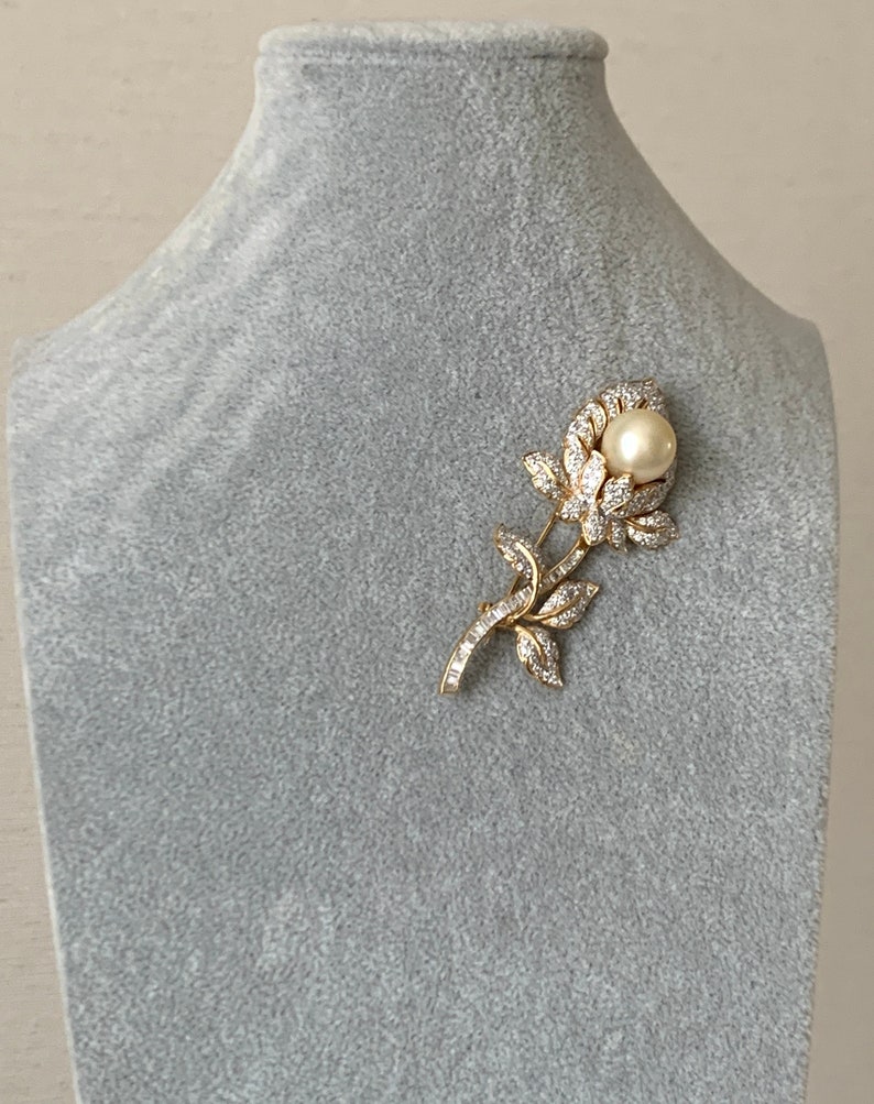 May include: A gold tone brooch in the shape of a rose with a single pearl in the centre. The rose is made of clear rhinestones.
