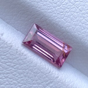 Op de afbeelding: Een rechthoekige, gefacetteerde edelsteen met een zachte roze tint. De edelsteen heeft een schoon, gepolijst uiterlijk, met scherpe randen en een subtiele interne structuur. Het licht reflecteert op de facetten, waardoor een spel van licht en schaduw ontstaat.