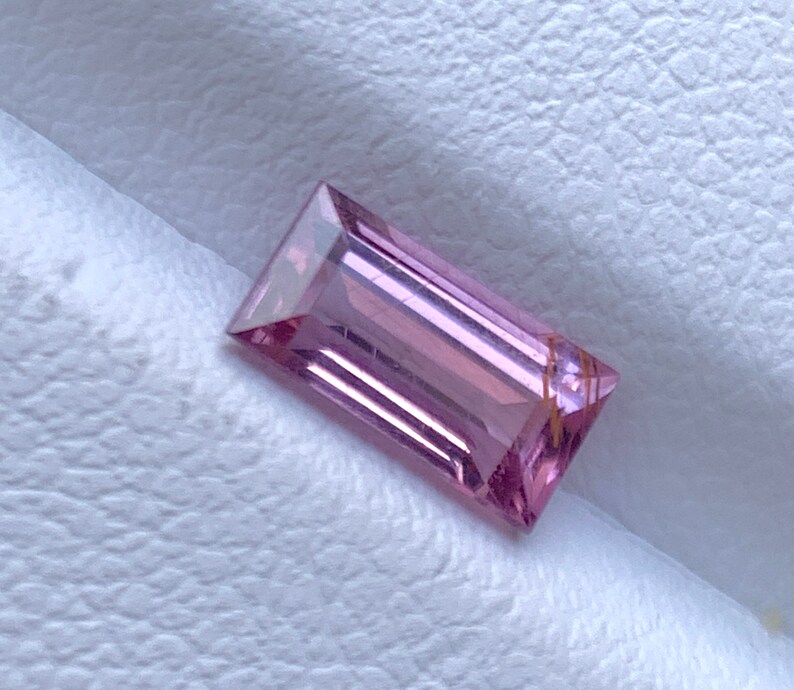 Op de afbeelding: Een rechthoekige, gefacetteerde edelsteen met een zachte roze tint. De edelsteen heeft een schoon, gepolijst uiterlijk, met scherpe randen en een subtiele interne structuur. Het licht reflecteert op de facetten, waardoor een spel van licht en schaduw ontstaat.