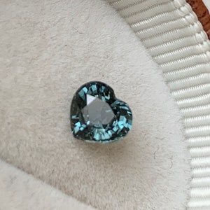 May include: A heart-shaped teal gemstone with facets, set in a white velvet ring box.