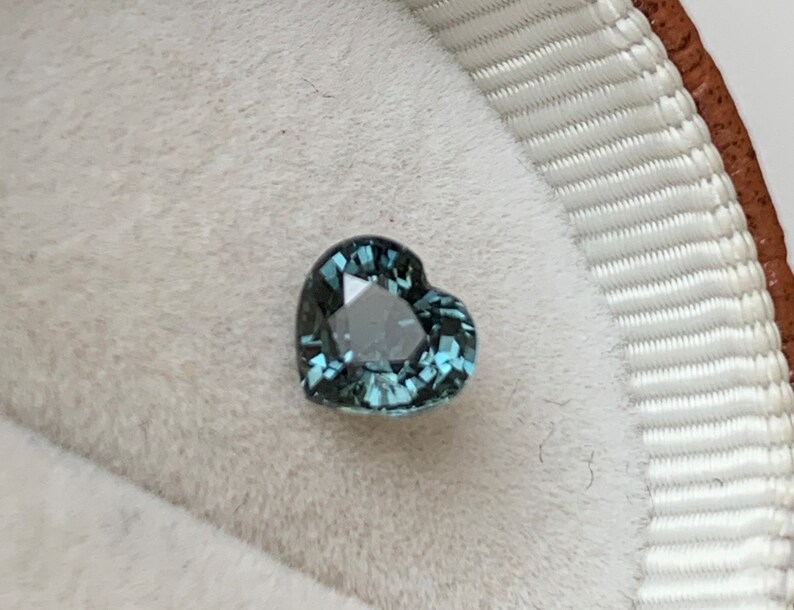 May include: A heart-shaped teal gemstone with facets, set in a white velvet ring box.
