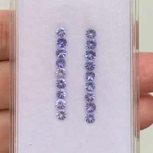 May include: Two rows of small, round, light purple gemstones in a clear plastic box with a white background.