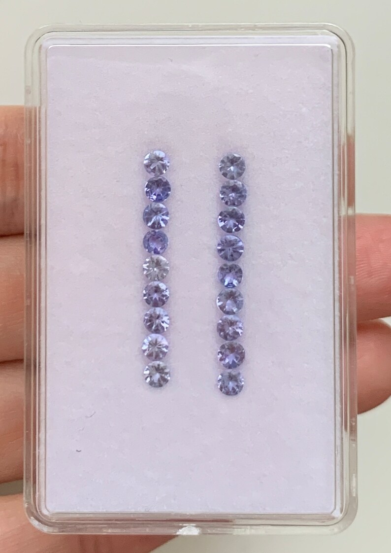 May include: Two rows of small, round, light purple gemstones in a clear plastic box with a white background.