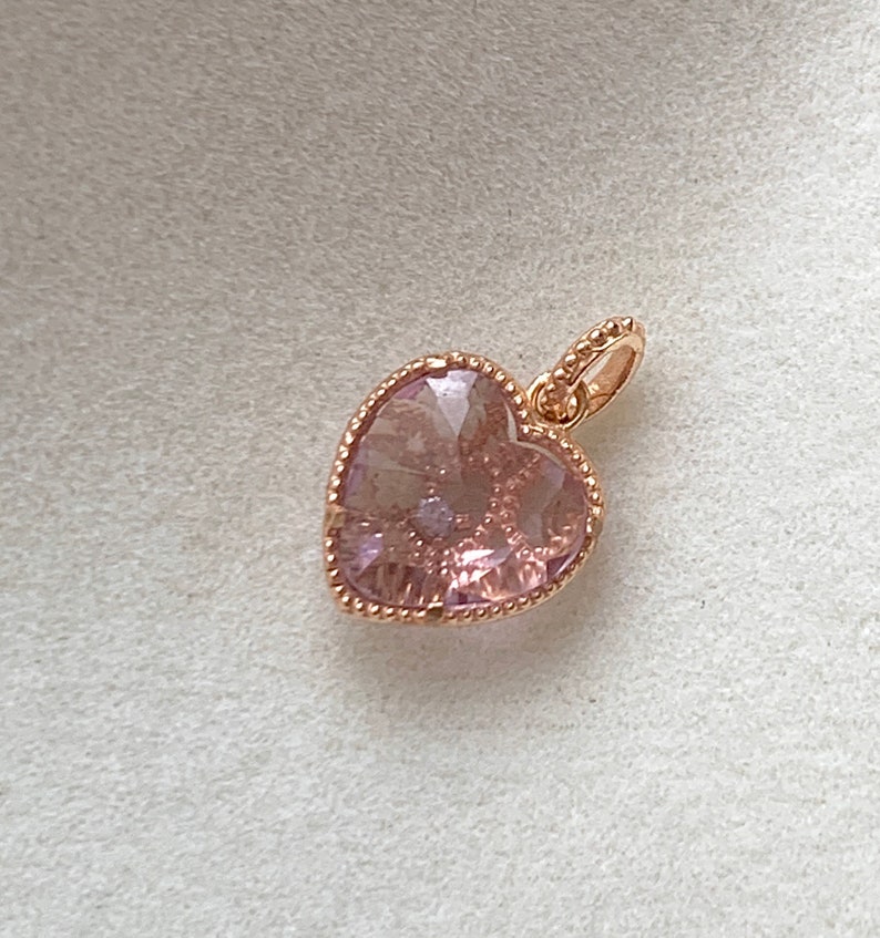 K&ouml;nnte beinhalten: Ein herzf&ouml;rmiger Anh&auml;nger aus Ros&eacute;gold mit einem rosa Kristall. Der Anh&auml;nger hat einen zarten Perlenrand.