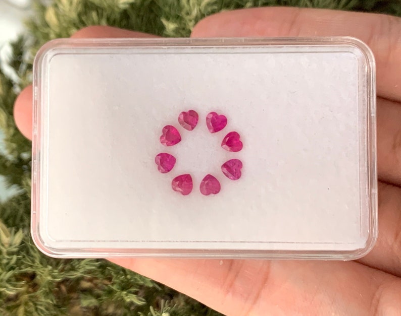 May include: A small white box with a clear lid containing a circle of ten pink heart-shaped gemstones.
