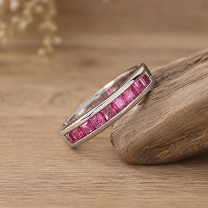 May include: A silver-toned band ring featuring a channel setting with a continuous row of square-cut, vibrant pink gemstones. The metal band has a polished finish and holds the stones securely in a straight line along the center of the ring.