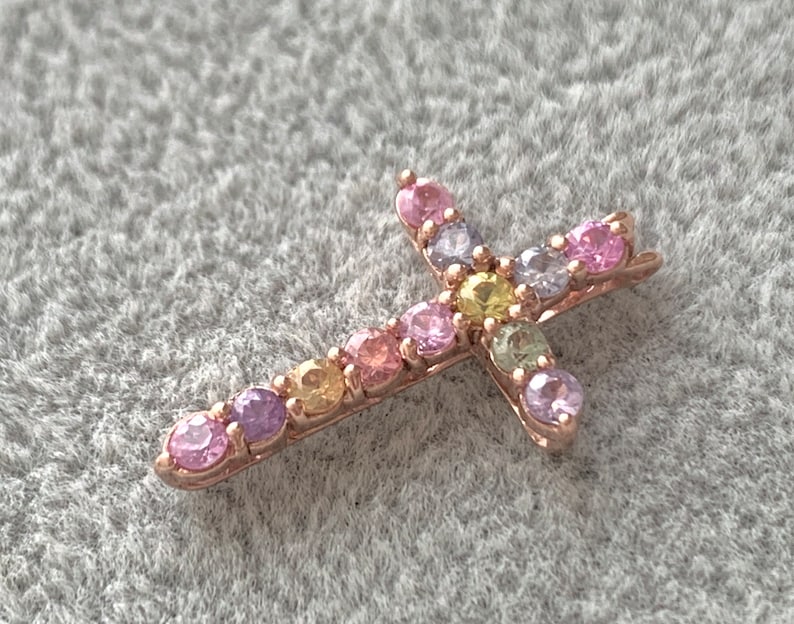 May include: A small gold cross pendant with colorful gemstones. The cross is adorned with pink, purple, yellow, and green gemstones.