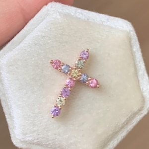 Könnte beinhalten: Ein roségoldener Kreuzanhänger mit bunten Edelsteinen. Das Kreuz ist mit einer Vielzahl pastellfarbener Steine besetzt, darunter Rosa, Lila, Blau und Grün. Der Anhänger wird auf einer weißen, samtartigen Oberfläche präsentiert.