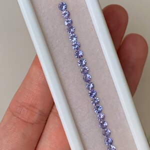 May include: A row of 17 round, light purple gemstones in a white display box.