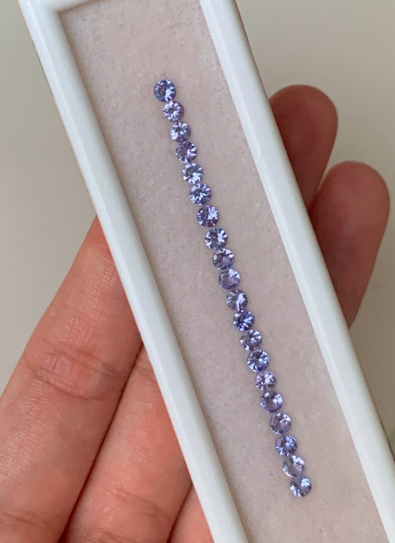 May include: A row of 17 round, light purple gemstones in a white display box.