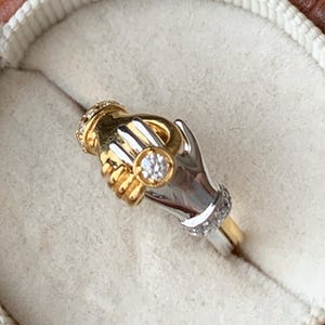 Könnte beinhalten: Ein zweifarbiger Gold- und Silberring mit zwei ineinander verschlungenen Händen und einem einzelnen Diamanten in der Mitte.