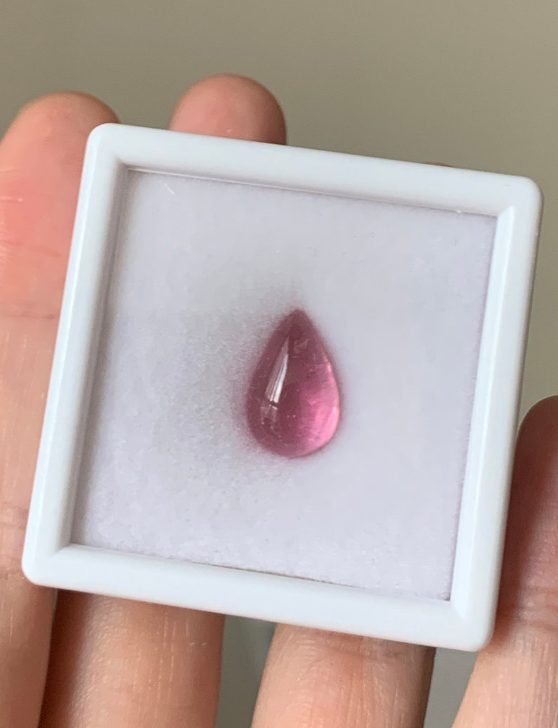 K&ouml;nnte beinhalten: Ein tropfenf&ouml;rmiger rosa Edelstein in einer wei&szlig;en Pr&auml;sentationsbox.