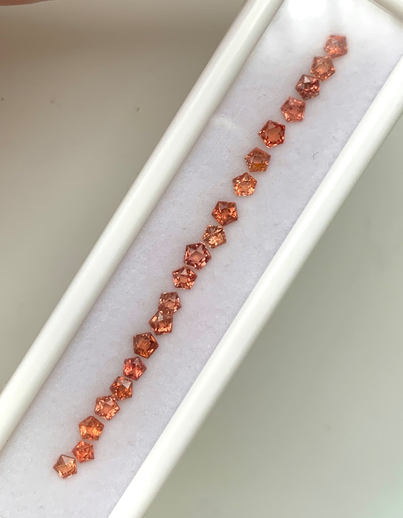 May include: A row of 15 orange and brown square-shaped gemstones in a white box.