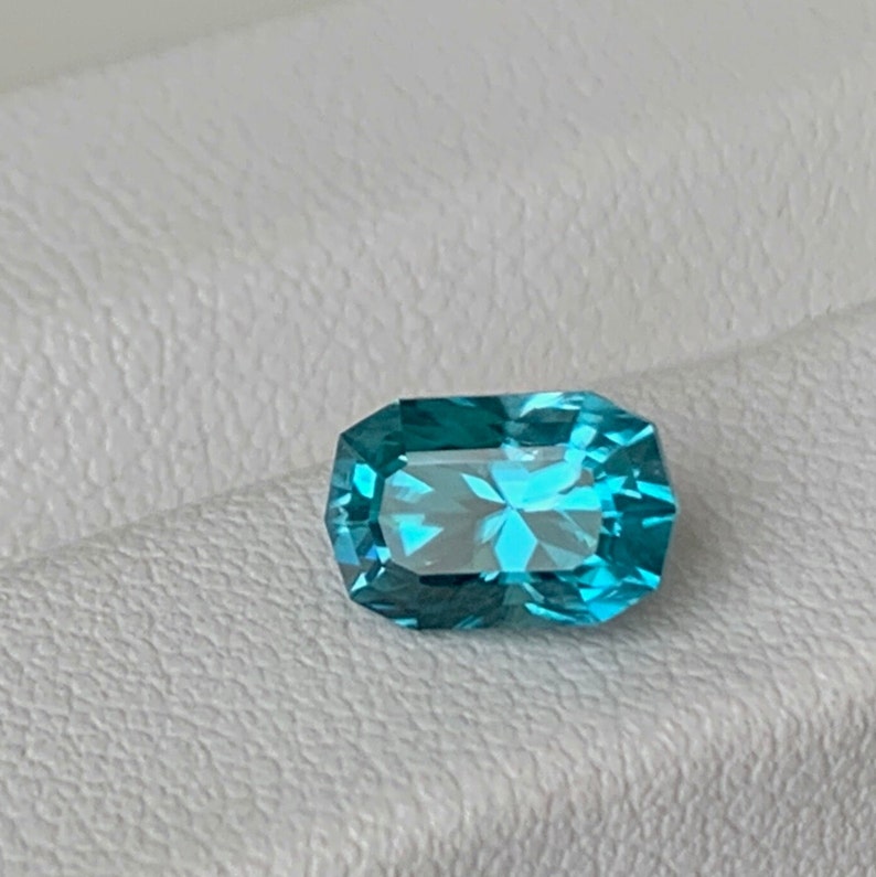 Puede incluir: Una gema azul vibrante con forma rectangular y corte facetado. La gema est&aacute; colocada sobre un fondo blanco.