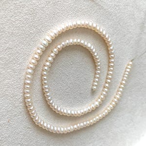Könnte beinhalten: Ein Strang kleiner, cremefarbener Perlen, spiralförmig angeordnet. Die Perlen sind gleichmäßig groß und geformt, wodurch ein zartes und elegantes Aussehen entsteht. Der Hintergrund ist eine helle, strukturierte Oberfläche, die den Glanz der Perlen hervorhebt.