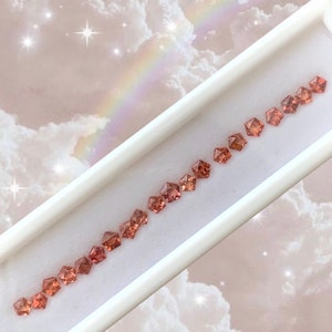 May include: A row of 15 small, faceted, orange-red gemstones in a white display case. The gemstones are arranged in a line, with the largest gemstone at the top and the smallest gemstone at the bottom.