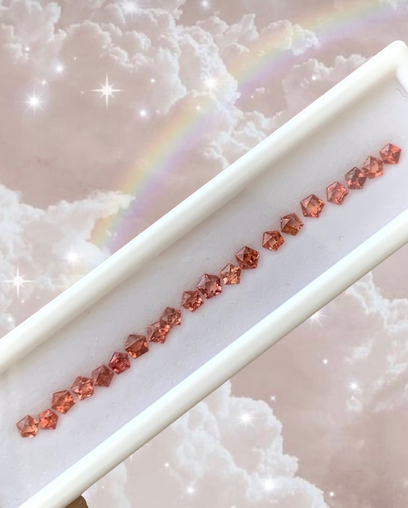 May include: A row of 15 small, faceted, orange-red gemstones in a white display case. The gemstones are arranged in a line, with the largest gemstone at the top and the smallest gemstone at the bottom.