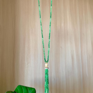 May include: A long, beaded necklace with varying shades of green beads. The necklace features a gold-colored pendant with a tassel of green beads. The necklace is displayed against a light brown wooden background.