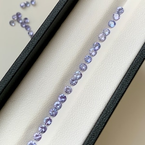 May include: A row of small, round, light purple gemstones arranged in a line on a white surface inside a black box.