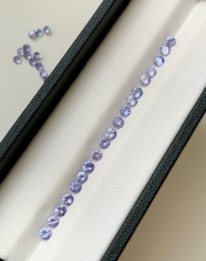 May include: A row of small, round, light purple gemstones arranged in a line on a white surface inside a black box.