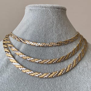 May include: Three beaded necklaces displayed on a gray mannequin. The necklaces feature a woven pattern of gold, silver, and black beads. The necklaces are arranged in a layered style, showcasing their intricate designs.