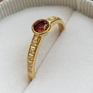 May include: A gold ring with a single red gemstone and a row of small diamonds on the band.