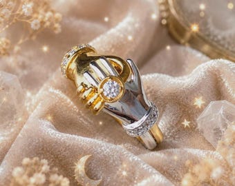 May include: A two-tone ring featuring two hands clasped together, crafted in polished gold and silver-toned metal. The design includes a central round clear gemstone set on one hand and small pavé-set stones along the bands of the ring.