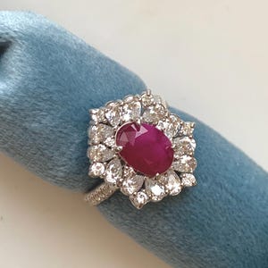 Könnte beinhalten: Ein silberner Ring mit einem großen, oval geschliffenen Rubin, umgeben von kleineren, klaren Edelsteinen. Der Ring ist vor einem blauen Hintergrund platziert, der das leuchtende Rot des Rubins und das Funkeln der Steine hervorhebt.
