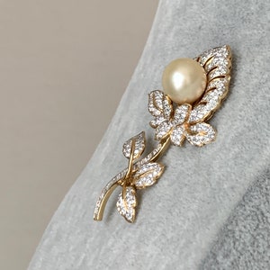 May include: A gold-tone brooch featuring a pearl and clear rhinestones in a floral design.