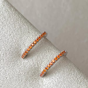 May include: A pair of silver hoop earrings with a thin band of orange gemstones.
