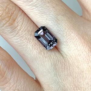 May include: A dark purple gemstone with a rectangular emerald cut. The gemstone is set in a ring and is being worn on a finger.