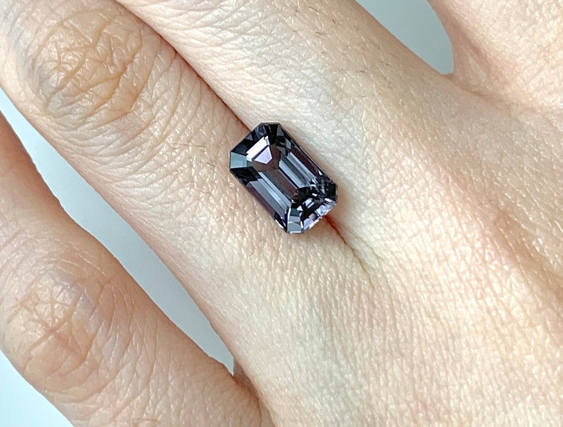 May include: A dark purple gemstone with a rectangular emerald cut. The gemstone is set in a ring and is being worn on a finger.