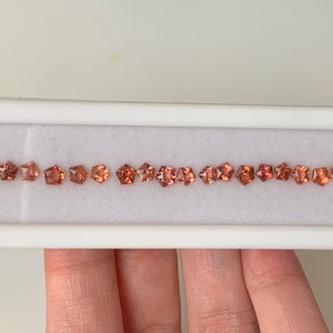 May include: A row of 20 small, square-shaped, orange-brown gemstones in a white display box.