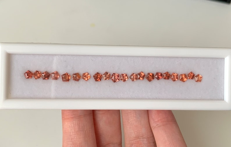 May include: A row of 20 small, square-shaped, orange-brown gemstones in a white display box.