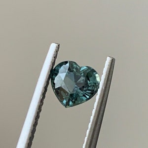May include: A heart-shaped green sapphire gemstone, held in tweezers.