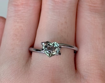 May include: A silver ring with a heart-shaped green gemstone. The ring is on a finger.