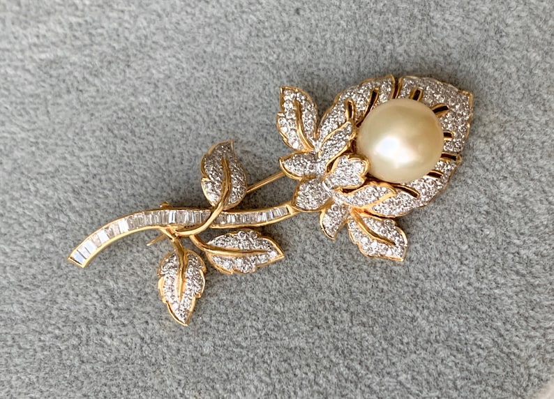 May include: A gold-tone brooch in the shape of a rose with a large pearl in the centre. The rose is made of small, clear stones and has a long stem with baguette-shaped stones.