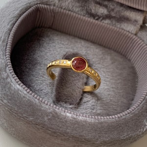 May include: A gold ring with a round, red gemstone surrounded by smaller, white gemstones. The ring is in a gray velvet ring box.