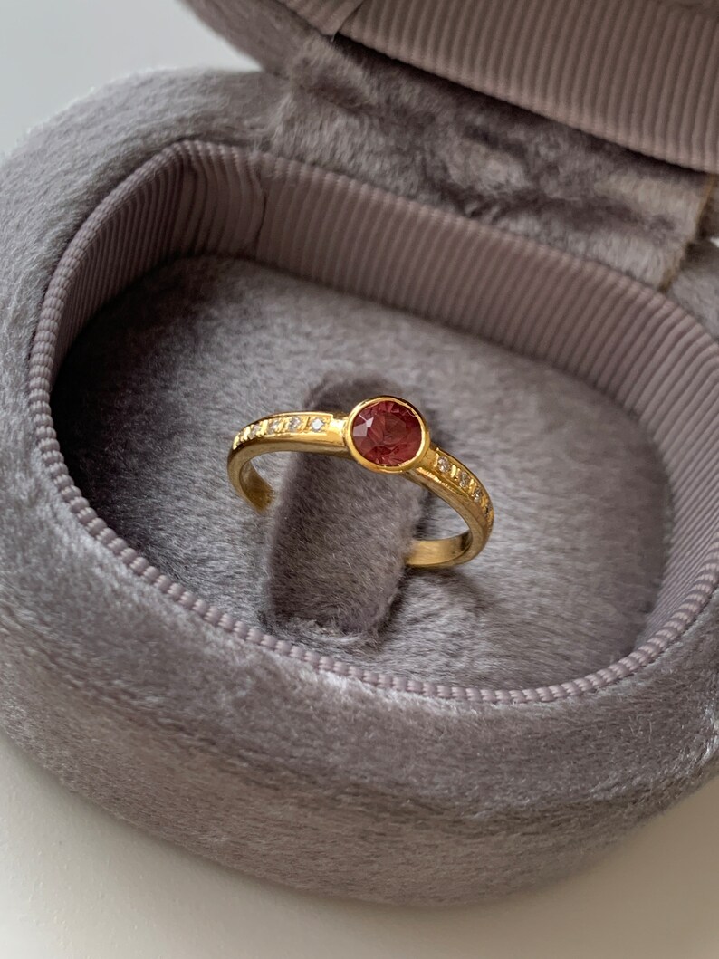 May include: A gold ring with a round, red gemstone surrounded by smaller, white gemstones. The ring is in a gray velvet ring box.