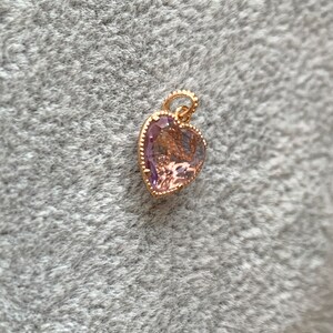 K&ouml;nnte beinhalten: Ein goldener herzf&ouml;rmiger Anh&auml;nger mit einem hellvioletten Edelstein. Der Anh&auml;nger hat einen detaillierten, verzierten Rand.