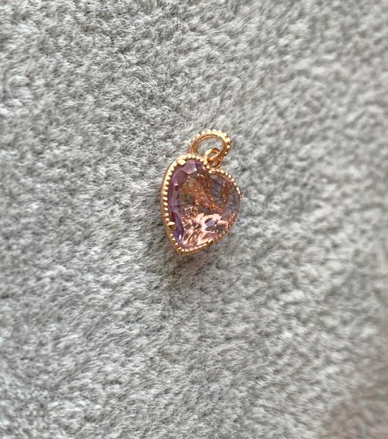 K&ouml;nnte beinhalten: Ein goldener herzf&ouml;rmiger Anh&auml;nger mit einem hellvioletten Edelstein. Der Anh&auml;nger hat einen detaillierten, verzierten Rand.