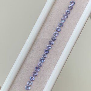 May include: A row of 20 small, round, light purple gemstones arranged in a line in a white box.