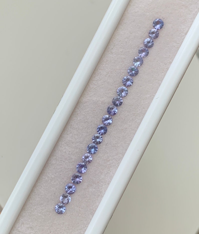 May include: A row of 20 small, round, light purple gemstones arranged in a line in a white box.
