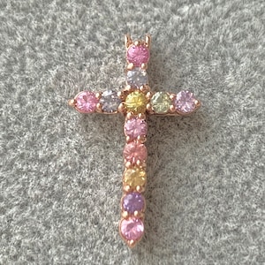 May include: A gold-tone cross pendant with a rainbow of colorful gemstones. The cross is adorned with small, round stones in shades of pink, purple, yellow, and light blue.