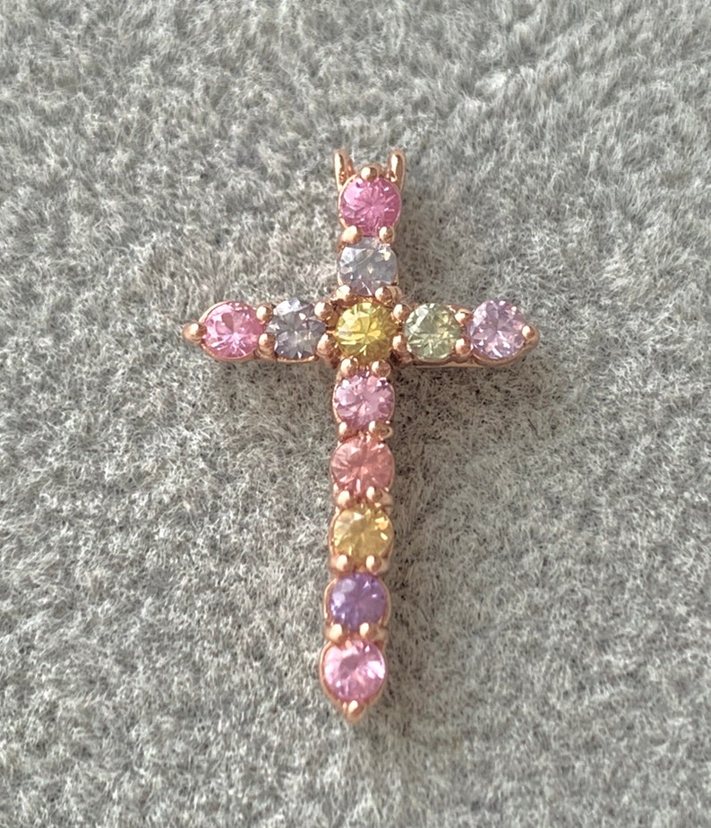 May include: A gold-tone cross pendant with a rainbow of colorful gemstones. The cross is adorned with small, round stones in shades of pink, purple, yellow, and light blue.