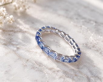 May include: A silver eternity band ring featuring a continuous row of baguette-cut blue sapphires set in a polished metal channel. The stones exhibit varying shades of light to medium blue, creating a uniform and elegant circular design.