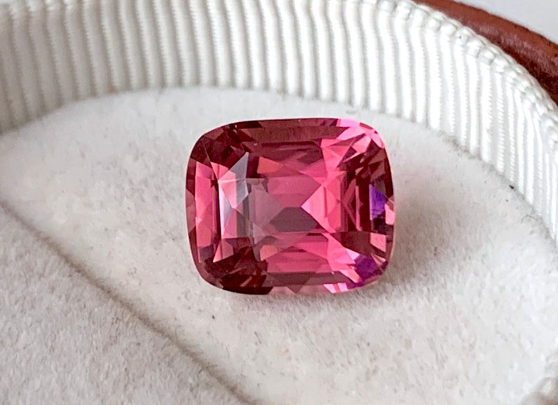 May include: A pink gemstone with a cushion cut, set in a white ring box.