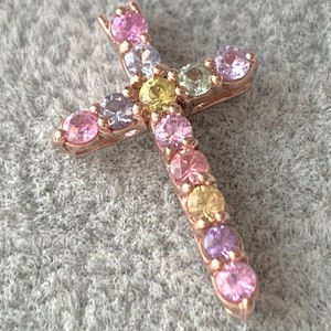May include: A small gold cross pendant with a variety of colorful gemstones. The gemstones are arranged in a pattern around the cross, creating a rainbow effect.