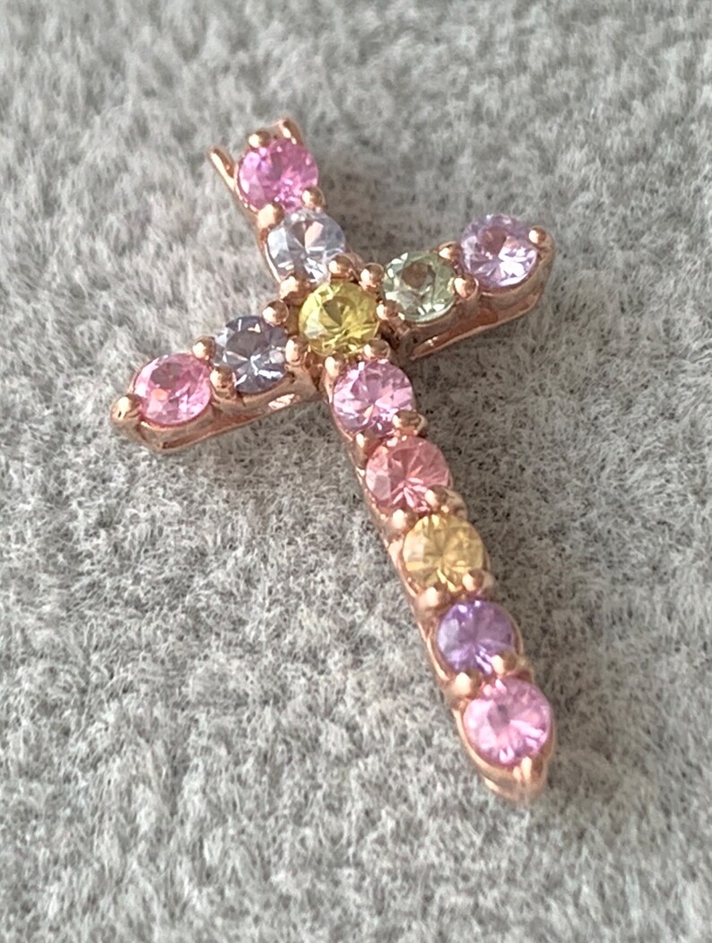 May include: A small gold cross pendant with a variety of colorful gemstones. The gemstones are arranged in a pattern around the cross, creating a rainbow effect.