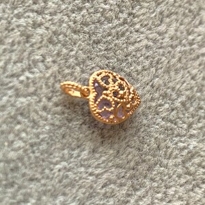 K&ouml;nnte beinhalten: Ein goldfarbener herzf&ouml;rmiger Anh&auml;nger mit einem filigranen Design und einem violetten Stein in der Mitte.
